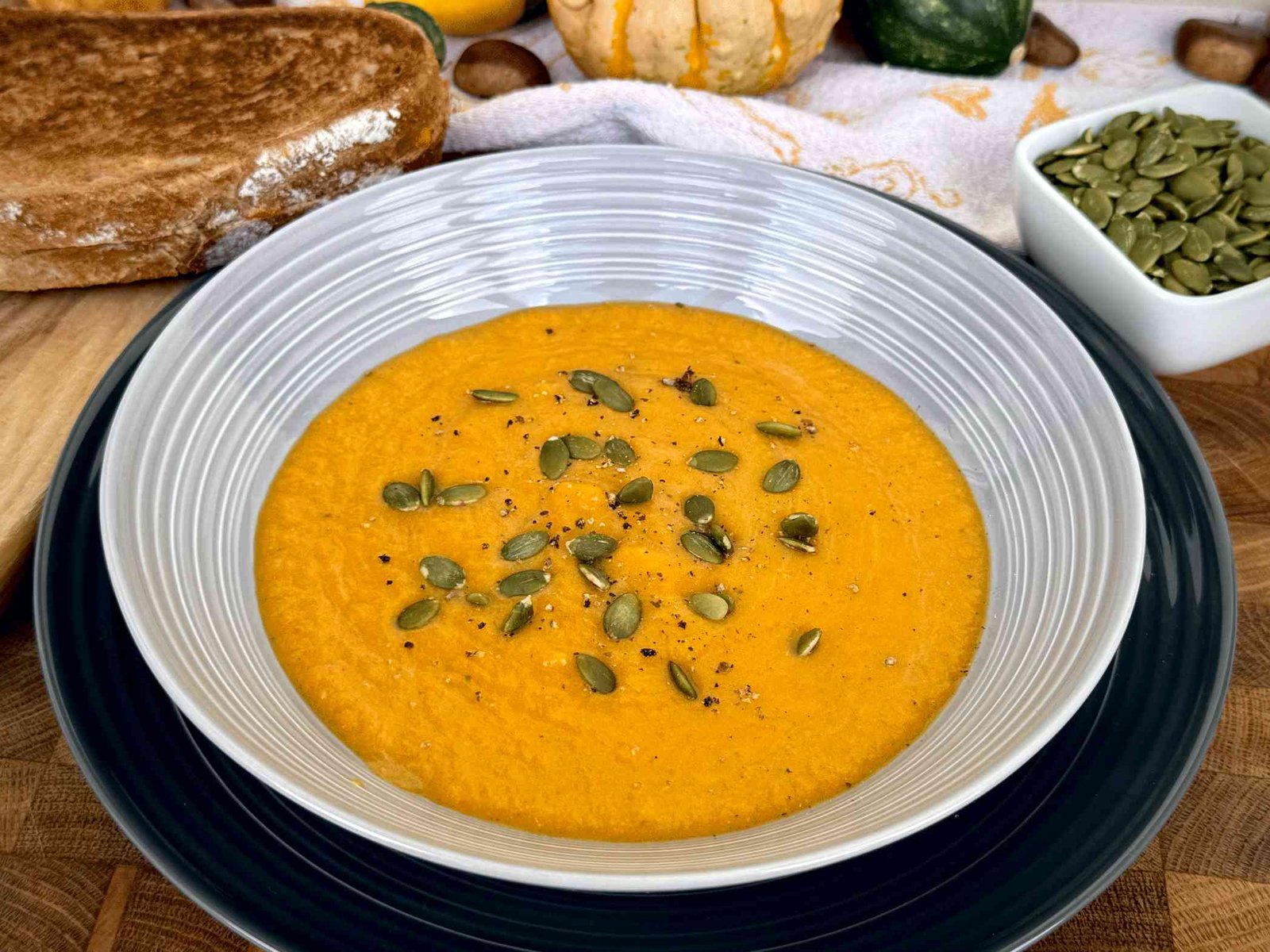 Krem supa od pečene bundeve u sivoj činiji, ukrašena sjemenkama golice, sa domaćim hljebom u pozadini.