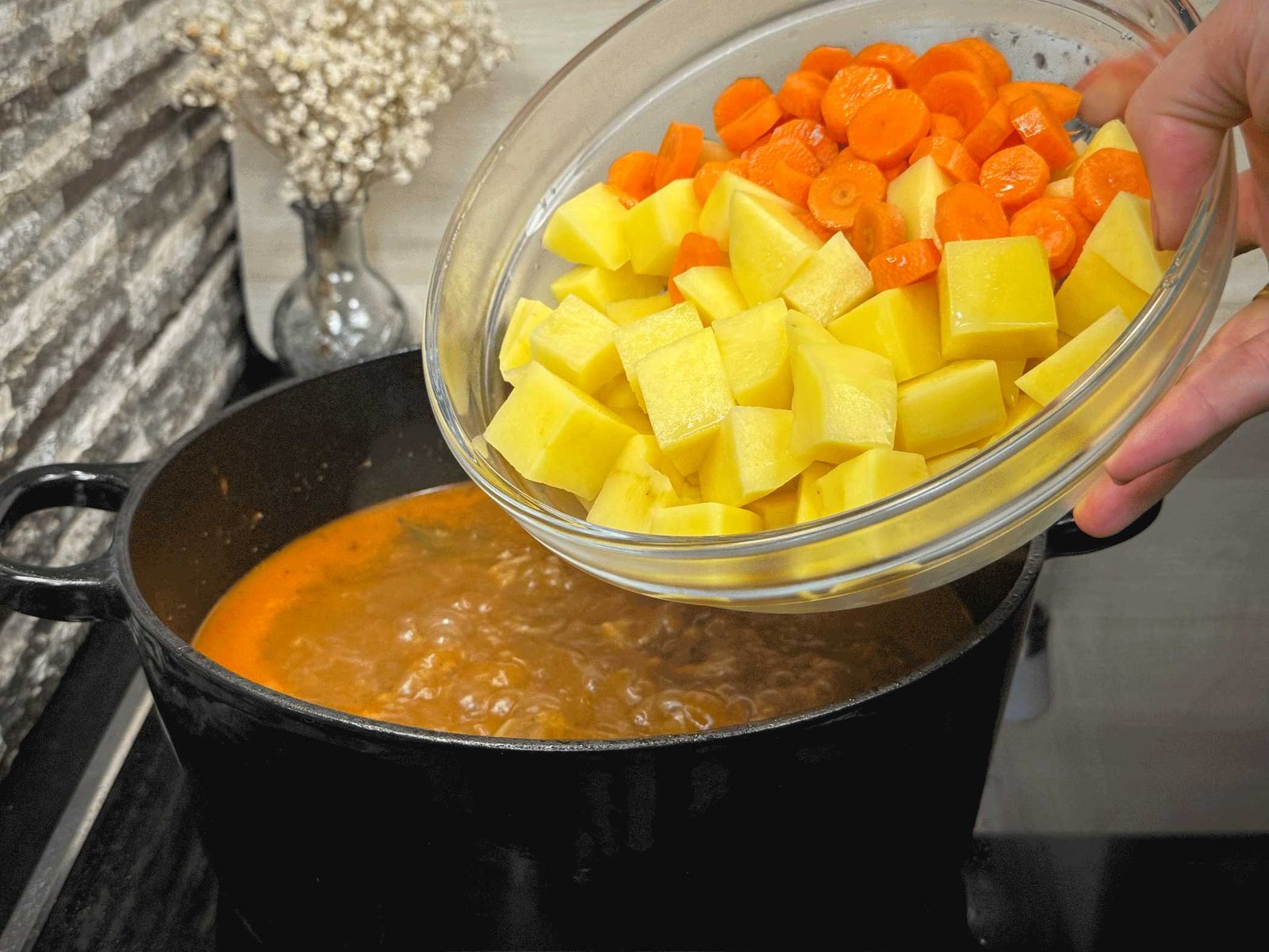 Ruka dodaje sjeckani krompir i kolutove mrkve iz staklene zdjele u lonac sa čorbom koja se kuha.