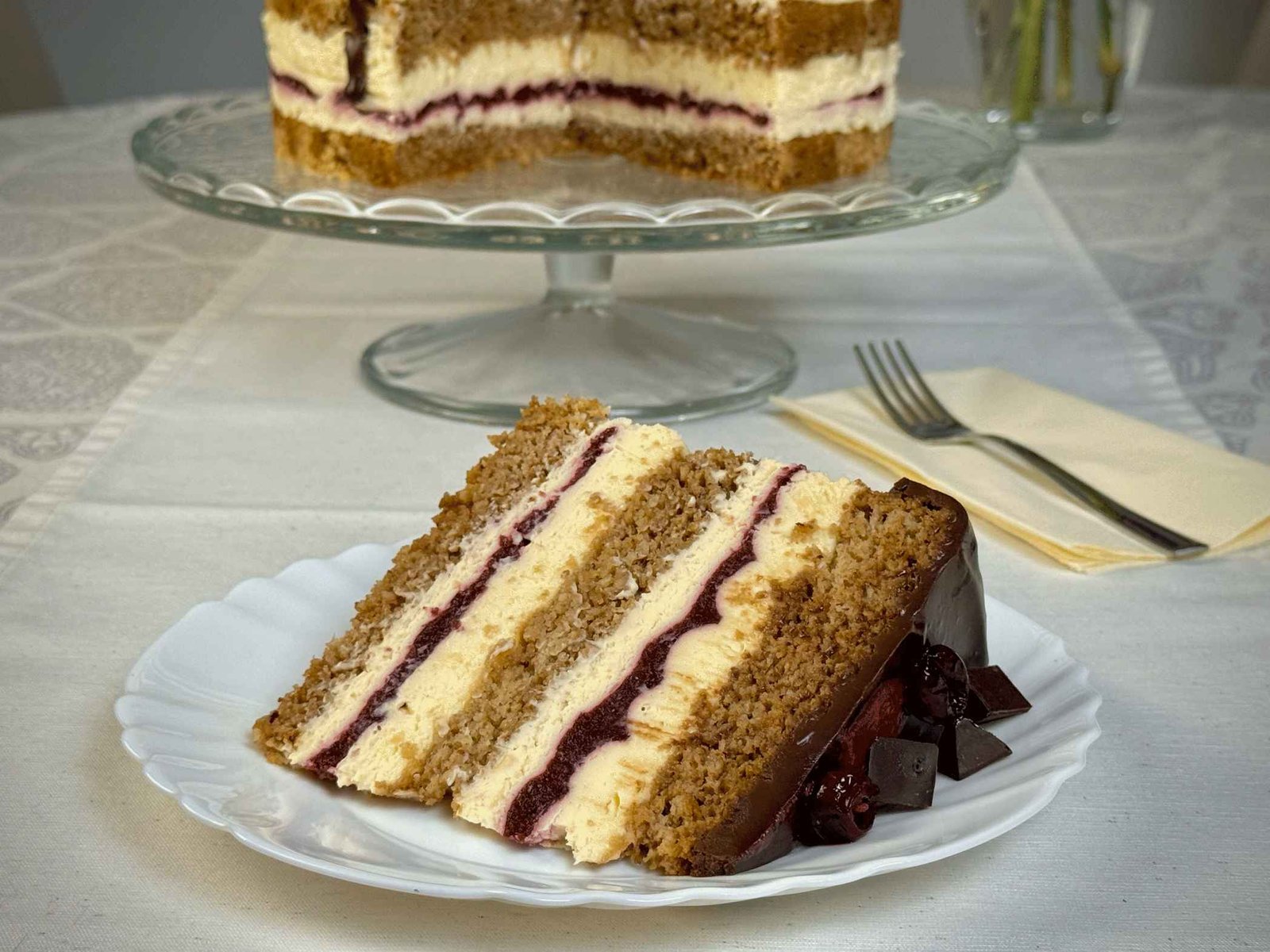 Komad torte sa višnjama na tanjiru u prvom planu, sa cijelom tortom u pozadini, naglašavajući slojevitost i bogatstvo deserta.