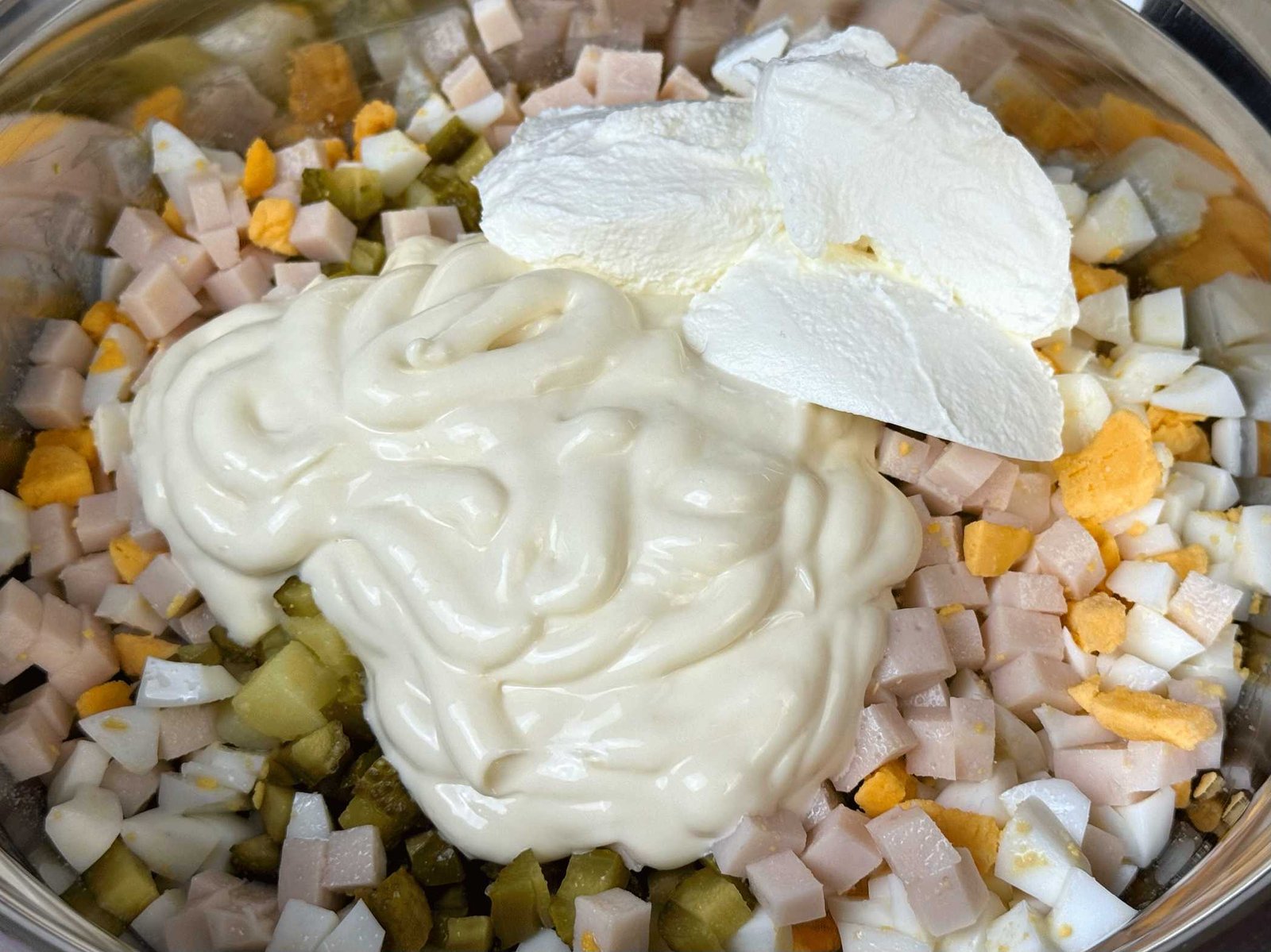 Dodavanje preliva od majoneze i kisele pavlake preko sjeckanih sastojaka za rusku salatu u velikoj metalnoj posudi.