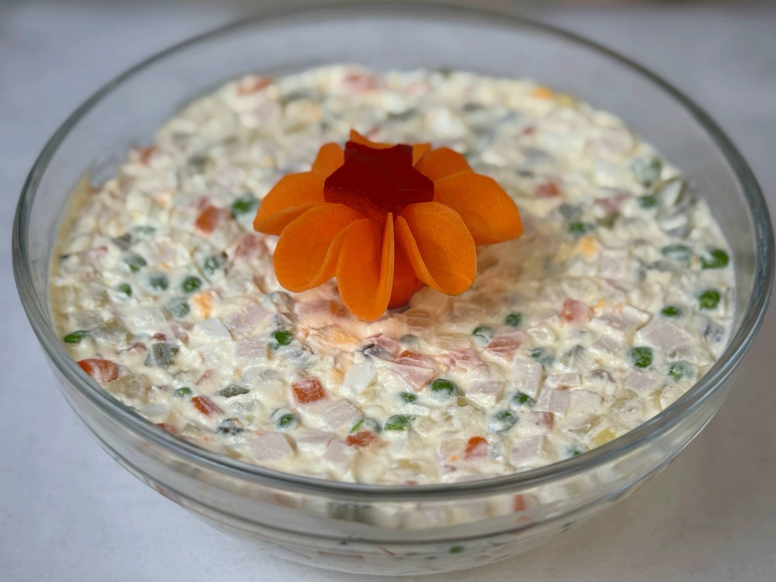 Kremasta ruska salata servirana u staklenoj činiji, sa savršeno sitno sjeckanim sastojcima i dekorativnim cvijetom od mrkve na vrhu.