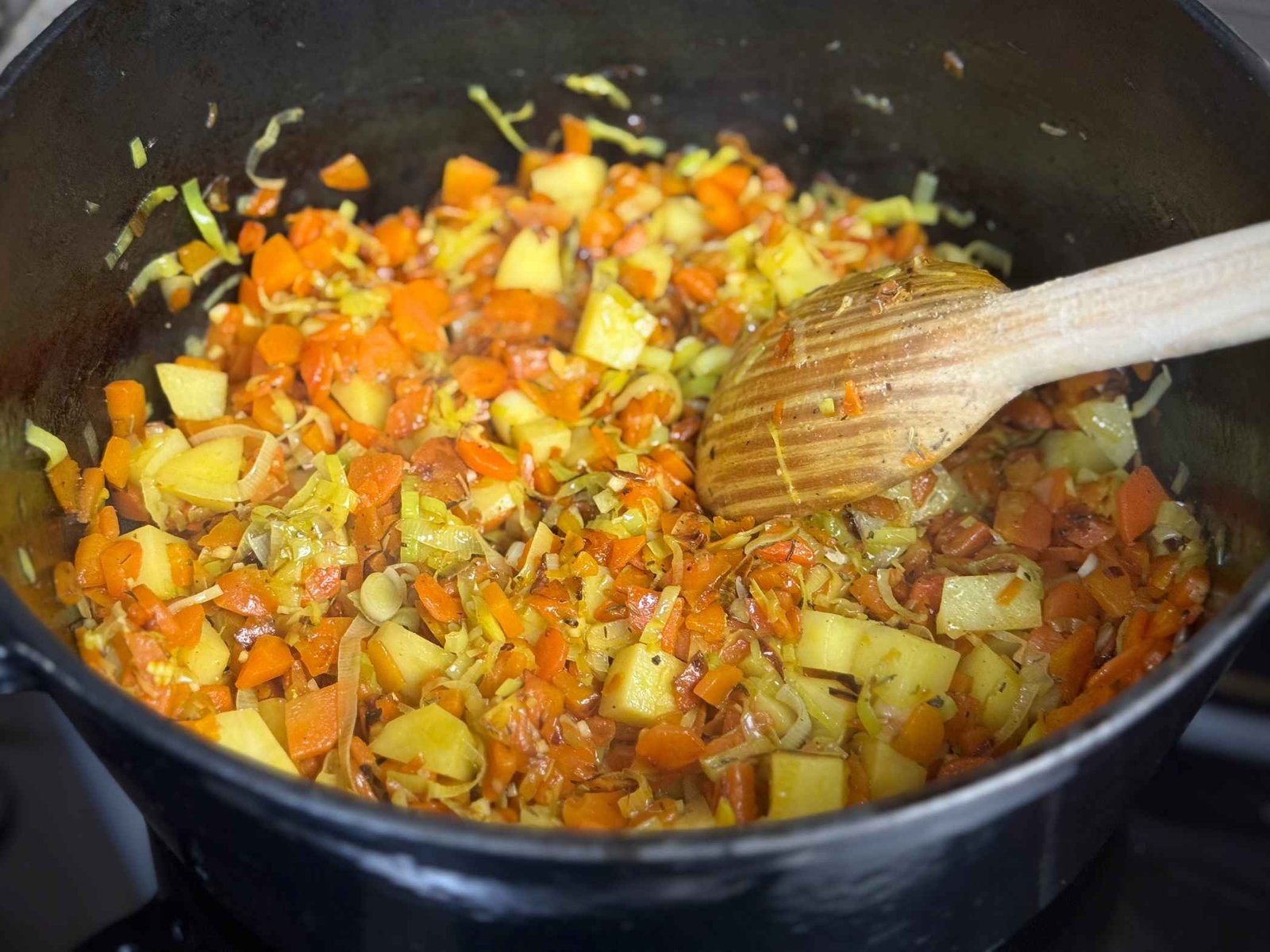 Dinstanje isjeckane mrkve, krompira i praziluka u crnoj šerpi uz miješanje drvenom kašikom.