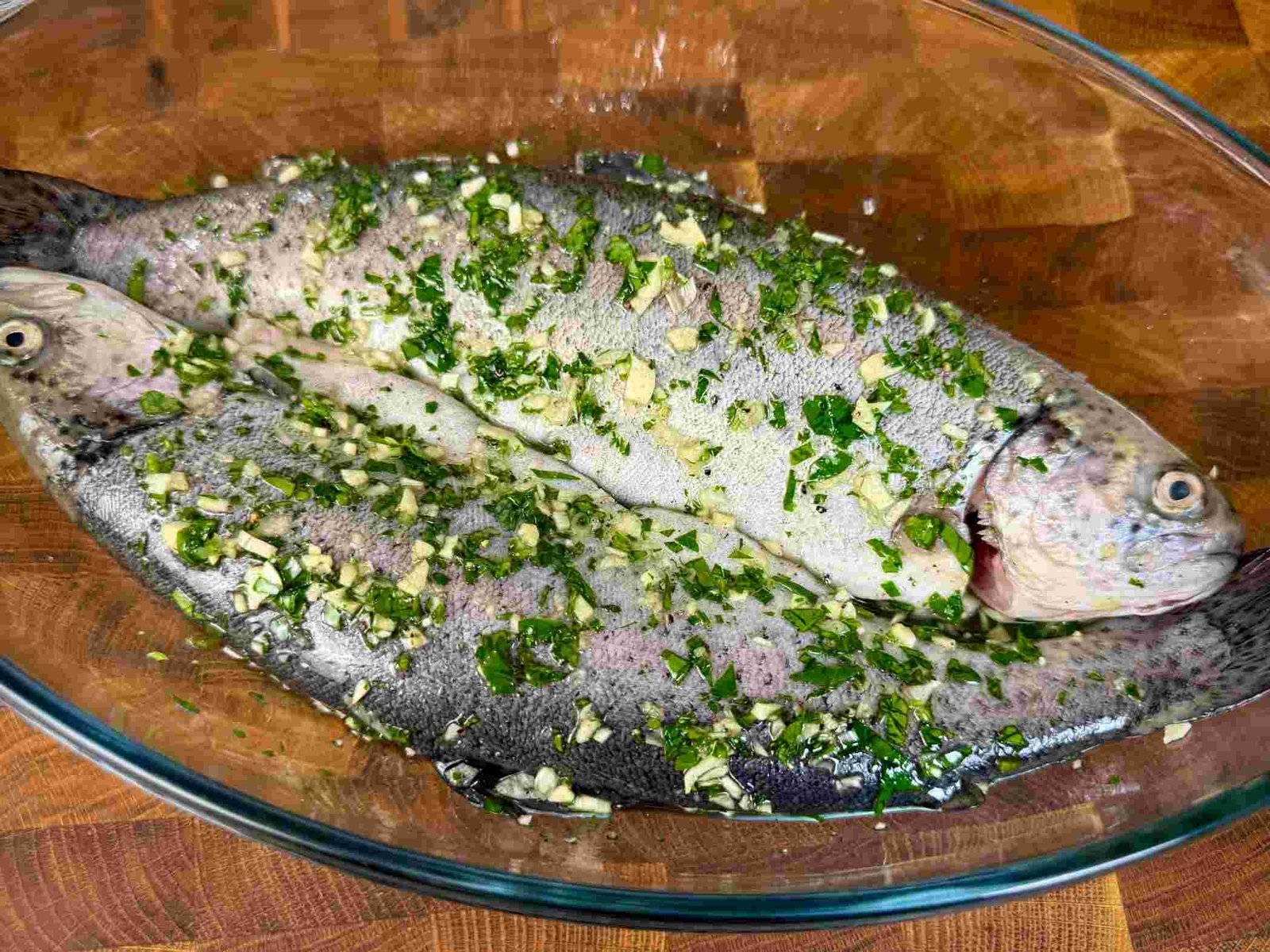 Dvije cijele pastrmke u staklenoj posudi prekrivene marinadom od bijelog luka, peršuna i ulja.