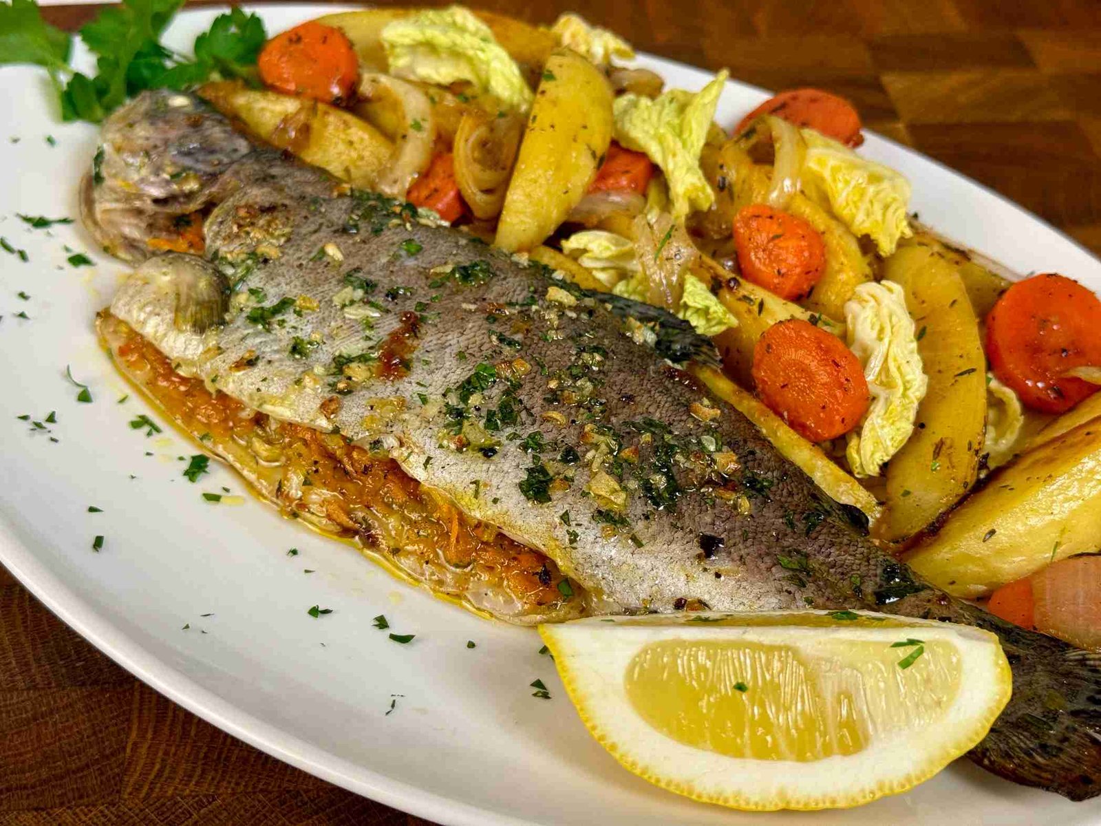 Cijela marinirana pastrmka pečena u rerni, poslužena sa pečenim krompirom, mrkvom i kriškom limuna. Jelo je posuto svježim peršunom i bijelim lukom.