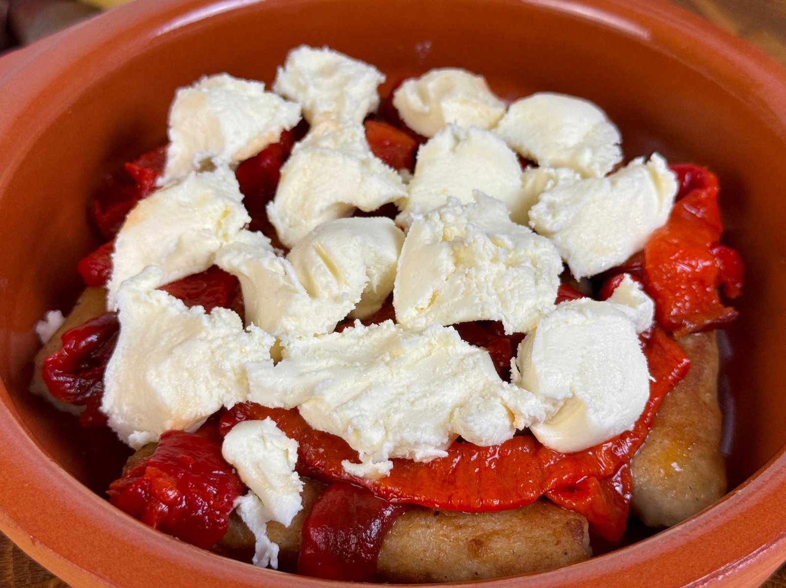 cevapi-sa-paprikama-i-kajmakom-prije-pecenja Ćevapi u keramičkoj posudi, prekriveni trakama pečene crvene paprike i komadićima kajmaka prije pečenja.
