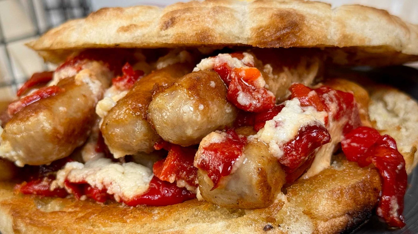 socni-cevapi-sa-kajmakom-u-hrskavoj-lepinji Krupni plan ćevapa sa rastopljenim kajmakom i pečenim crvenim paprikama u hrskavoj lepinji.